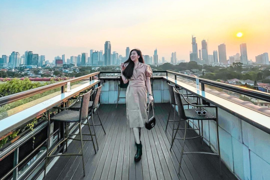 A woman walks along the rooftop terrace of La Vue Bar at The Hermitage Jakarta during sunset, city skyline in the background.