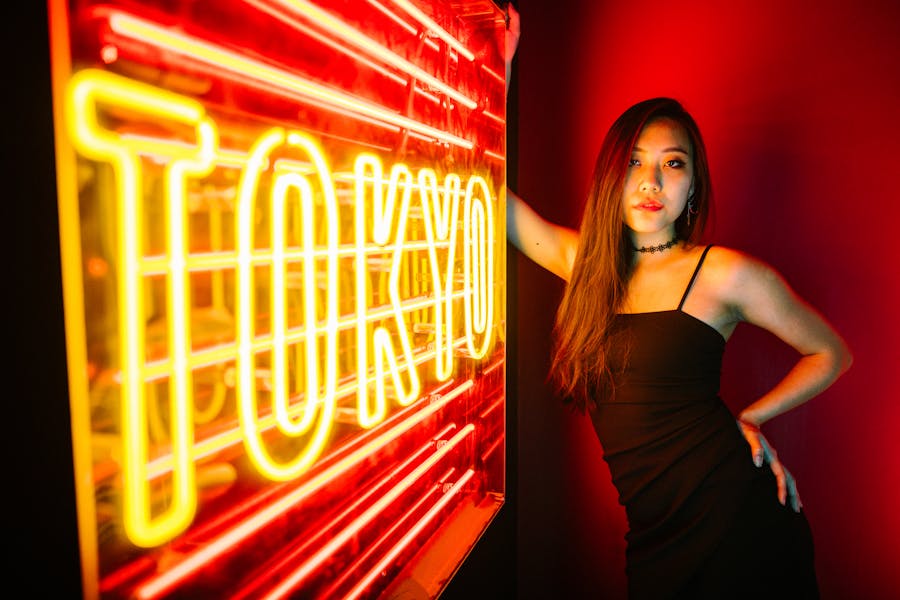 A ladyboy wearing a black dress poses indoors next to a bright yellow and red Tokyo neon sign on a dark background.