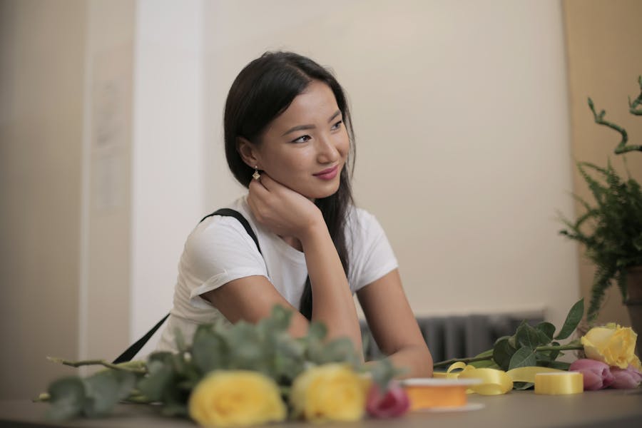 Ladyboy sitting at a table in a cozy café with flowers, looking away thoughtfully in a calm indoor London setting.